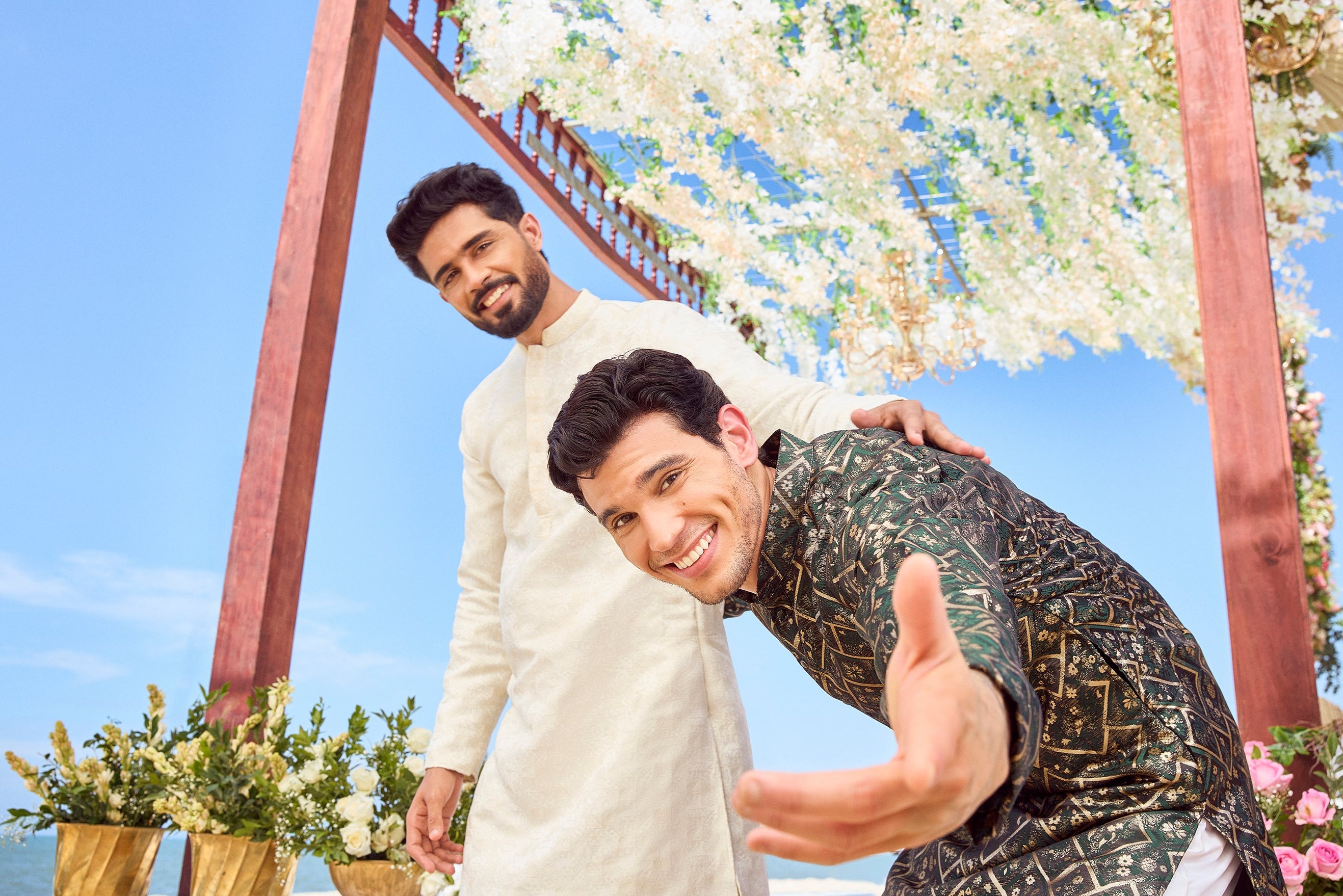 Two men posing under a floral arch on a beach with clear blue sky.