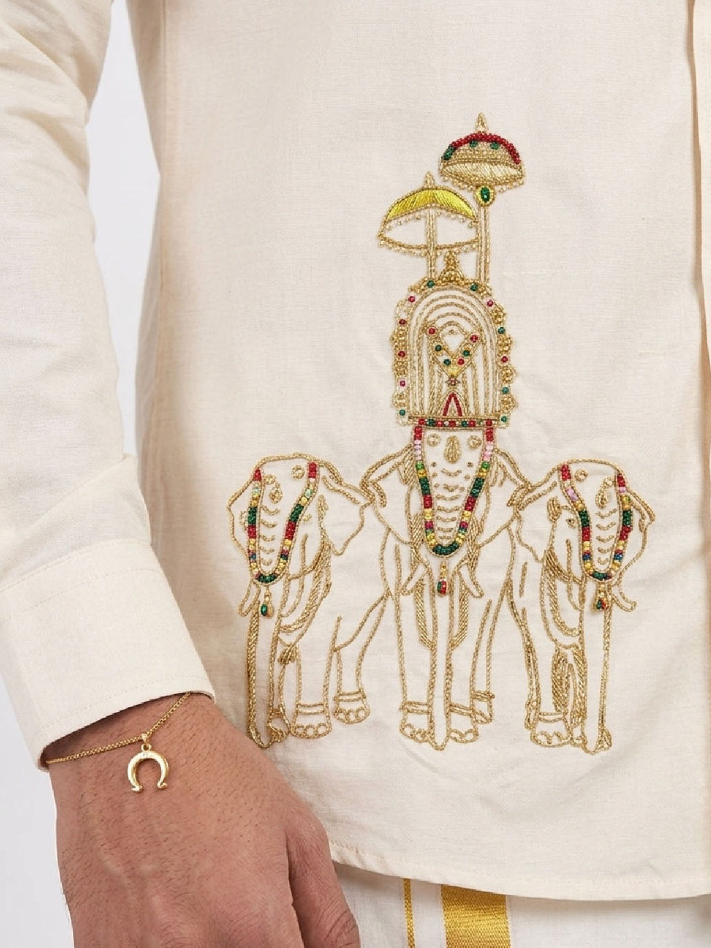 Close-up of a cream-colored kurta featuring an intricate gold embroidered design of three elephants carrying a decorated chariot, with a model's hand wearing a gold bracelet visible on the left.